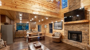 A spacious living area with a wooden floor, a fireplace, a dining table, and a large TV mounted on the wall. - New! “WaterTastic” Private Cabin heated Indoor Pool, Theater, fantastic view  (Gatlinburg)