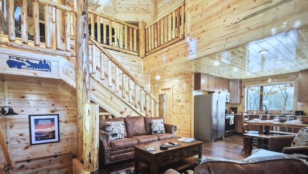 A spacious wooden cabin with a central staircase, a cozy living area with a sofa and coffee table, and a kitchen area with a refrigerator and sink. - New! “WaterTastic” Private Cabin heated Indoor Pool, Theater, fantastic view  (Gatlinburg)