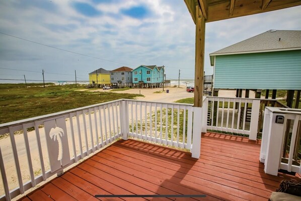 A wooden deck with a white railing, overlooking a beach with colorful houses and a clear sky.