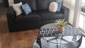 Ein schwarzes Ledersofa mit gemusterten Kissen, ein Glastisch mit einer gewebten Unterlage und eine Topfpflanze.