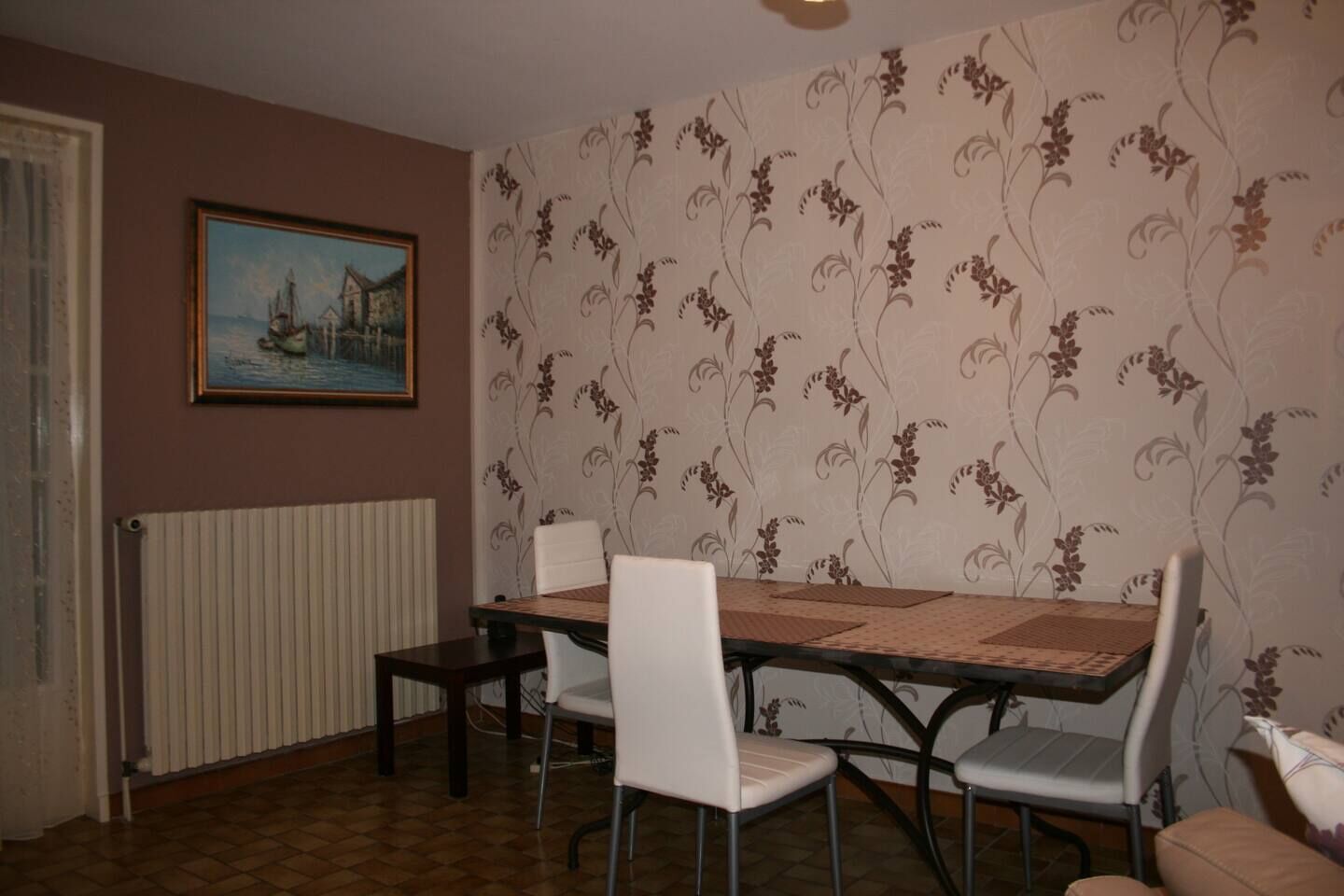A dining area with a table and chairs, a radiator, a floral wallpaper, and a painting on the wall.