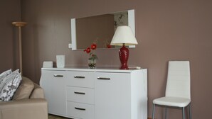 Et hvidt skab med skuffer, en rød vase med blomster, en hvid stol og et spejl over skabet.