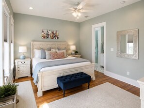 A bedroom with a bed, bedside tables, a ceiling fan, a large mirror, and a door leading to another room.