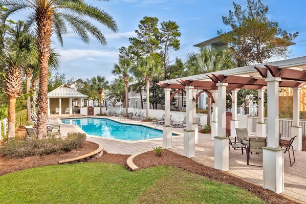 A backyard with a swimming pool, palm trees, and a covered patio area with chairs.