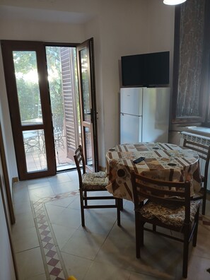 A dining area with a table covered in a patterned tablecloth, surrounded by wooden chairs with cushioned seats. A refrigerator is visible in the background, and a television is mounted on the wall.