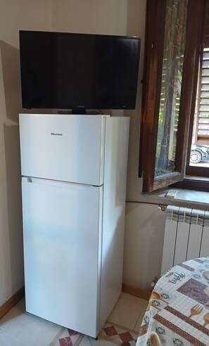 A compact kitchen with a white refrigerator, a flat-screen TV mounted above it, and a window with a view of greenery outside. - Petit Chateau - Room with kitchen (Montecatini Terme)