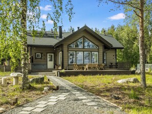 Mökki, jossa on suuri kuisti, katettu terassi ja soralla päällystetty polku, joka johtaa sisäänkäynnille.