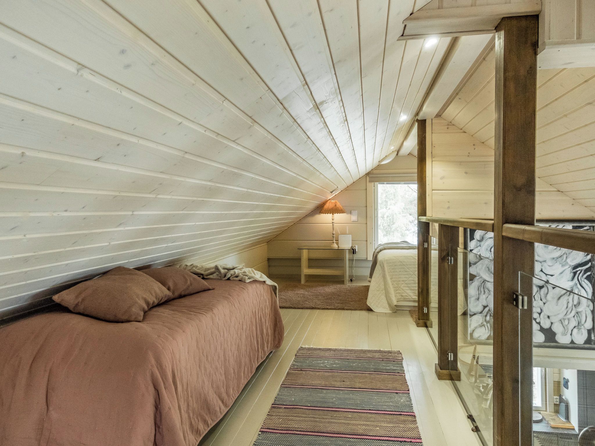 A bedroom with a sloped ceiling, a bed with a brown cover, a small table with a lamp, and a view through a glass door.