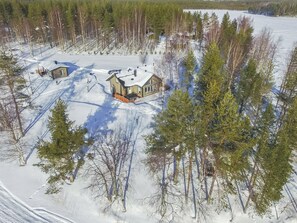 Lumipeitteinen maisema, jossa on kaksi puutaloa ja niiden ympärillä puita.
