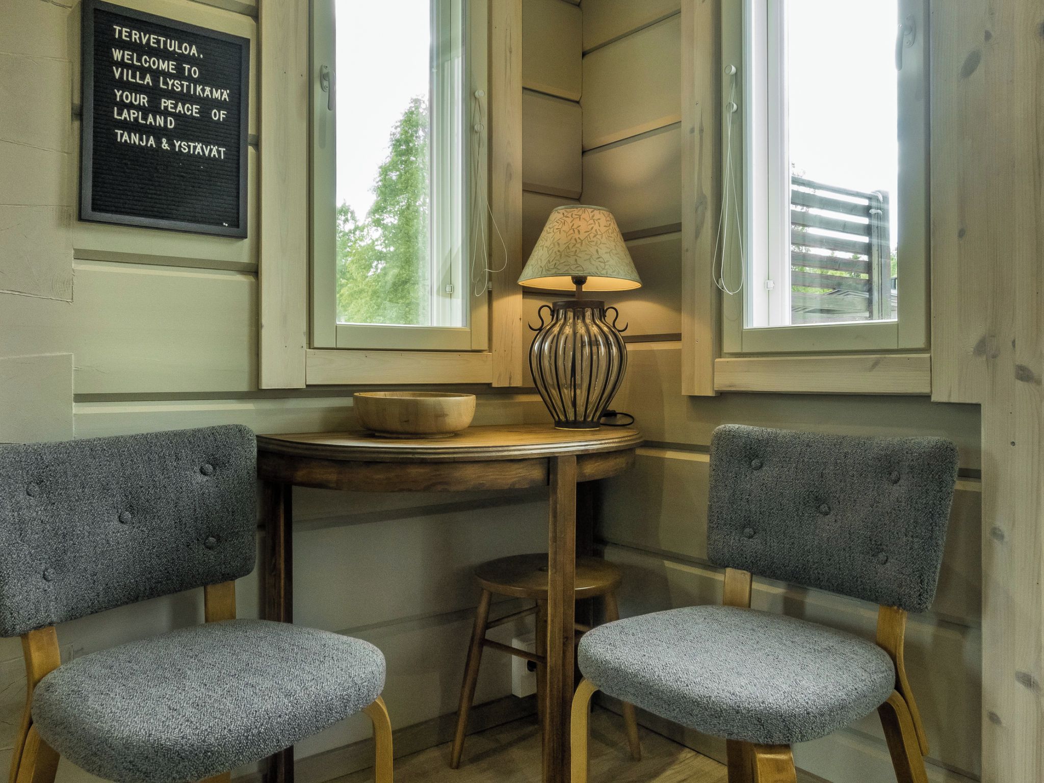A corner room with two grey upholstered chairs, a small round table with a bowl, a lamp, and a framed welcome sign.