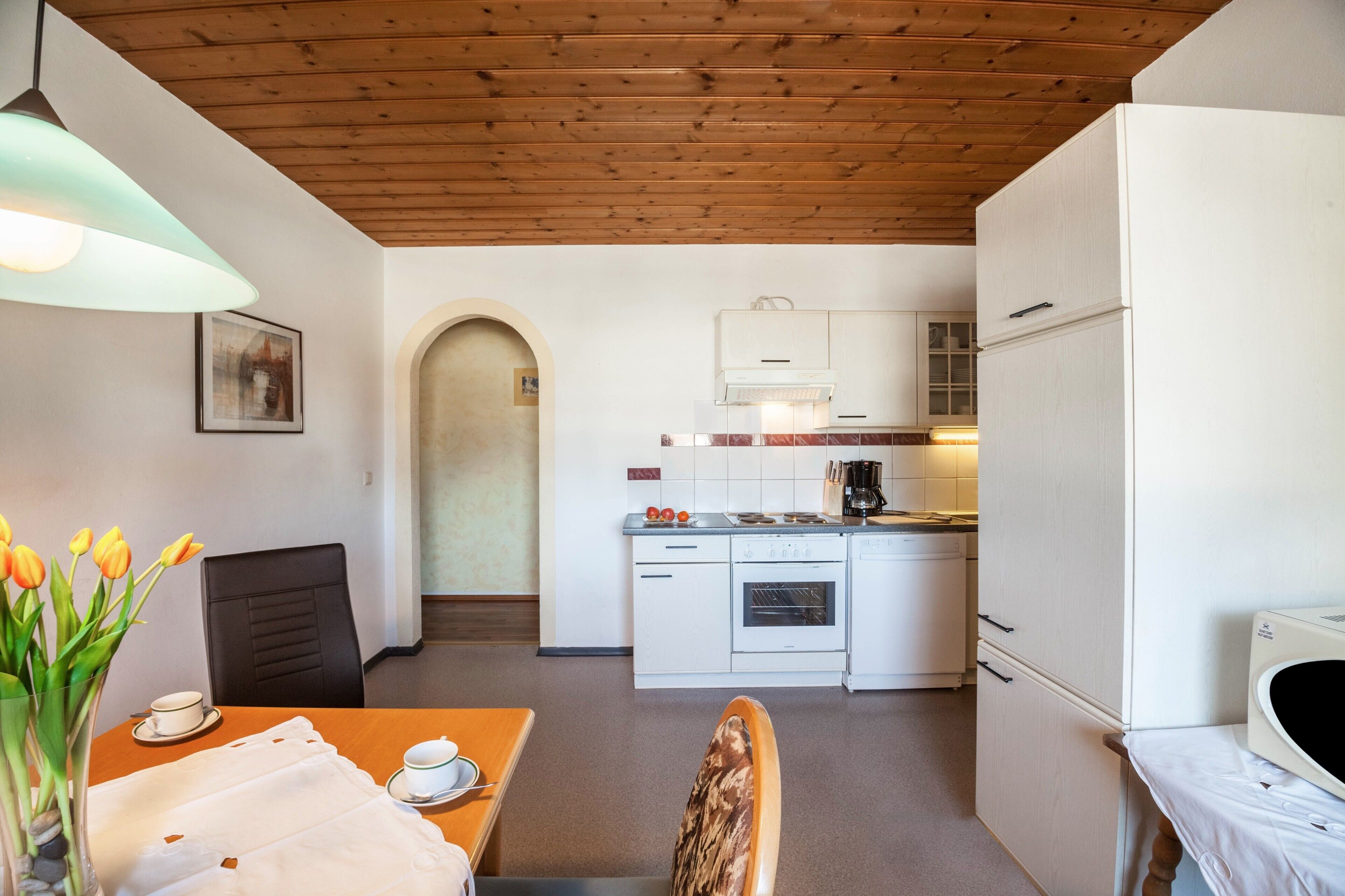 Une cuisine compacte avec des meubles blancs, une cuisinière et un four à micro-ondes. On y trouve un coin repas avec une table et des chaises, ainsi qu'un vase de tulipes posé sur la table.