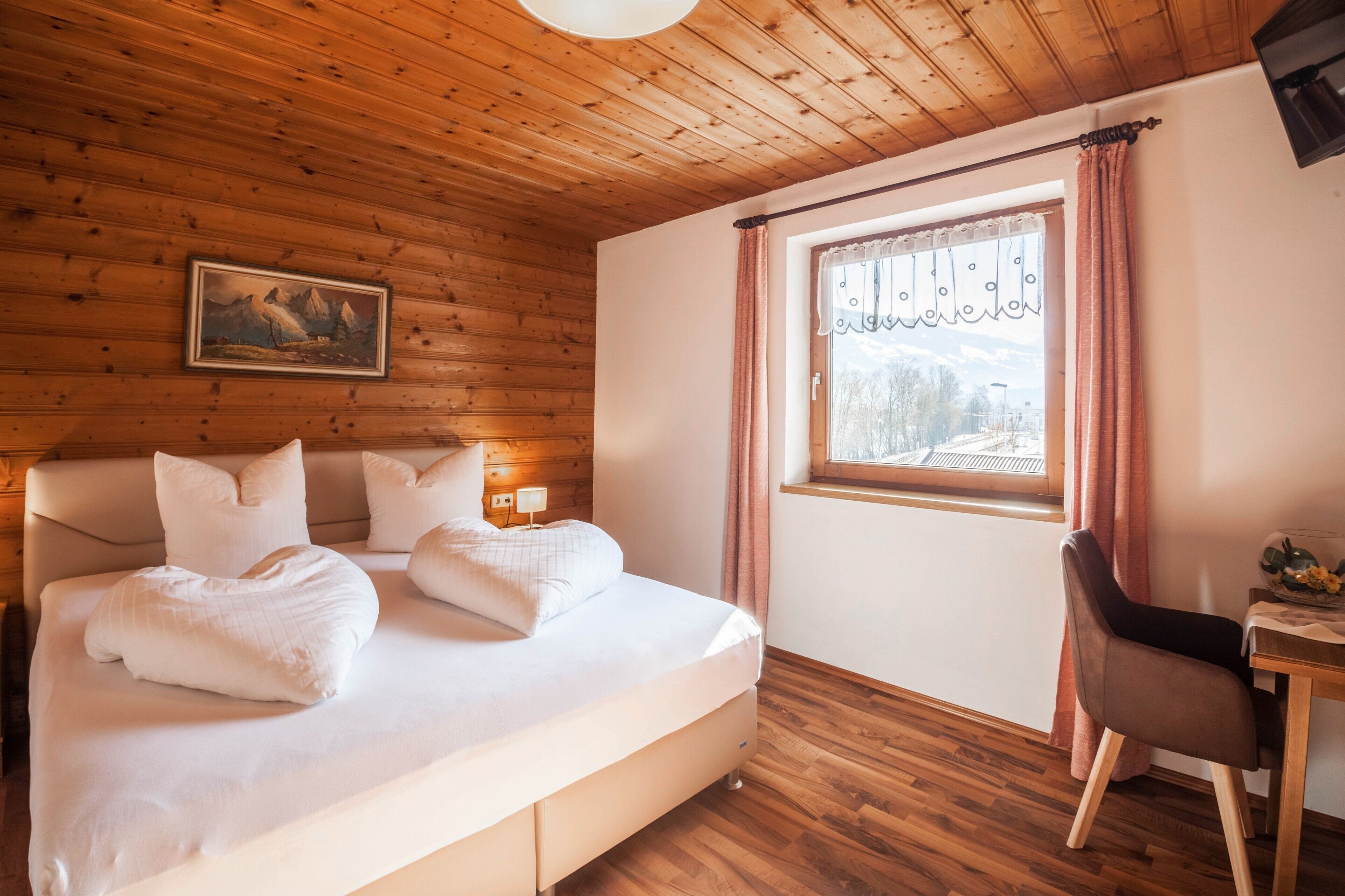 A wooden-paneled room with a bed, a window with curtains, a chair, and a painting on the wall.