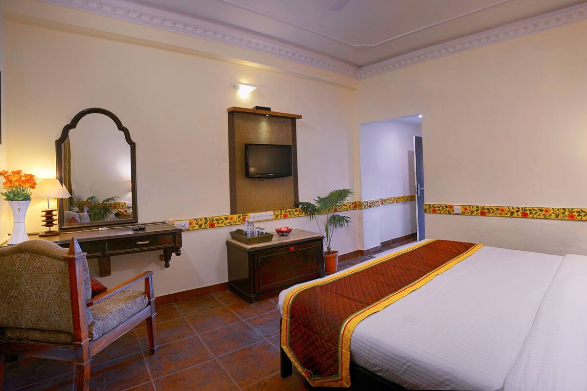 A hotel room with a bed, a desk with a chair, a mirror, a television, and a small table with a plant.