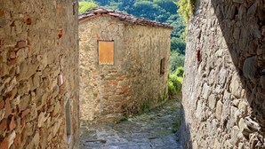 Un camí de pedra estret que condueix a un edifici rústic amb teulada de teules, envoltat de murs de pedra i un paisatge de muntanyes al fons.