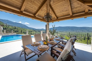 An outdoor dining area with a view of mountains, a pool, and a pergola with a hanging light fixture.