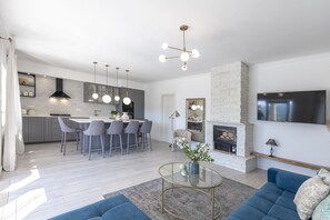 A modern kitchen with grey cabinets, a central island with bar stools, a fireplace, and a flat-screen TV mounted on the wall.