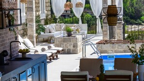 A sunlit outdoor seating area with stone walls, white curtains, wicker lamps, and a pool. - Villa Velaga (Gruda)