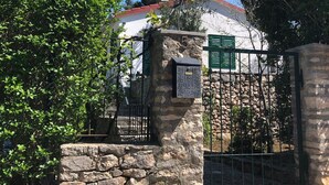 A stone wall with a metal gate, a stone building with a red roof, and a tree in the background. - Holiday Home Pavarotti (Supetar)