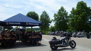A group of people dining under a PAULANER tent at an outdoor event with motorcycles parked in the background.