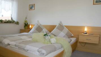 Een net opgemaakt bed met een geruit kussen, een nachtkastje met een lamp en een raam met gordijnen.