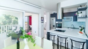 A modern kitchen with white countertops, a stainless steel range hood, and a built-in microwave. There is a dining area with a white table and chairs, and a balcony with outdoor seating. - Apartments Villa Bella Vista (Orebic)