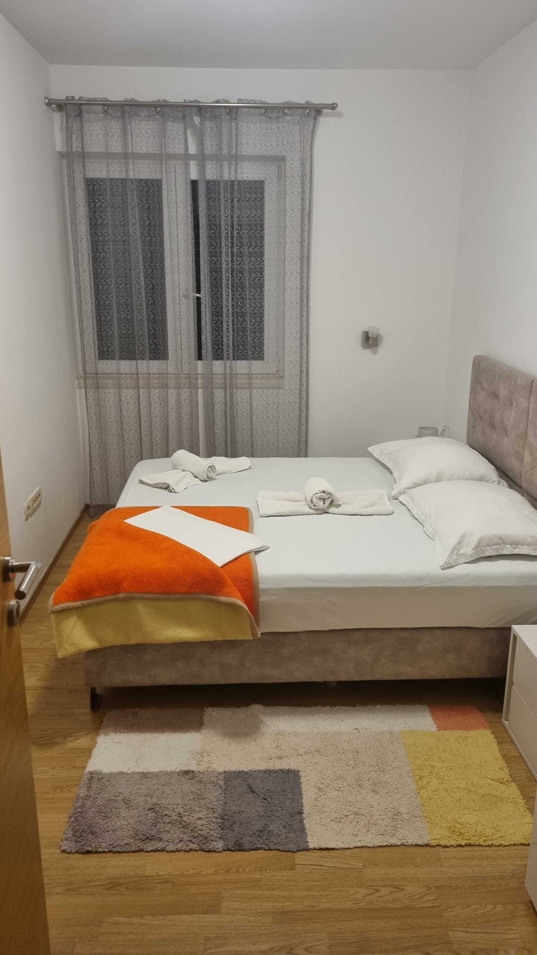 Une chambre à coucher avec un lit, une fenêtre ornée de rideaux légers, une table de chevet et un petit tapis.