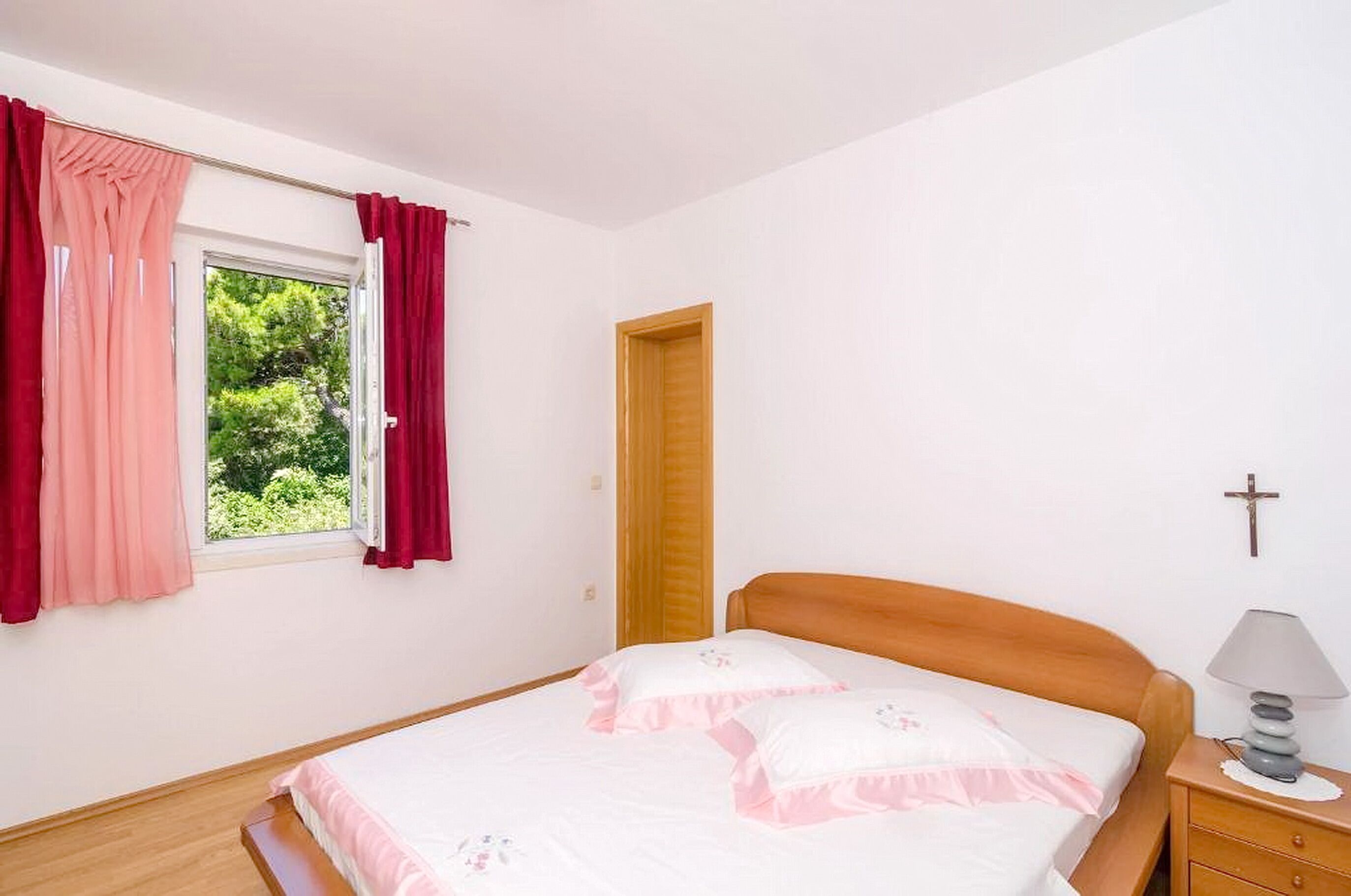 A bedroom with a bed, a nightstand with a lamp, a wooden door, and a window with curtains.