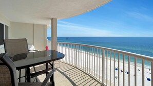 Ein Balkon mit Tisch und Stühlen, der auf einen Strand mit weißer Brüstung und blauem Himmel blickt.