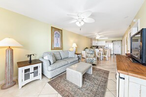 A living room with a sofa, coffee table, and a TV stand. There is a ceiling fan, a lamp, and a painting on the wall.