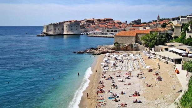 A coastal town with a historic fortification, a sandy beach with umbrellas, and clear blue waters.