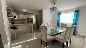 A dining area with a glass table and four chairs, a chandelier, a kitchen with white cabinets, and a window with blue curtains. - Inviting 3-bedroom Apt in Santo Domingo! (Santo Domingo Norte)