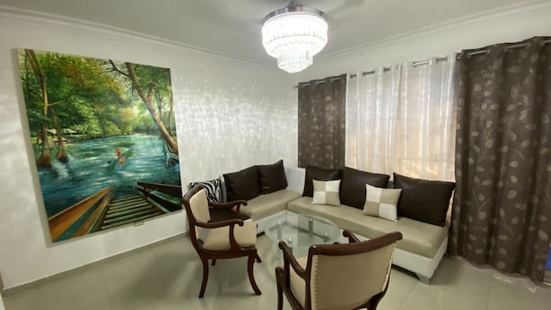 A living room with a sofa, two armchairs, a glass coffee table, a chandelier, a painting of a river scene, and patterned curtains.