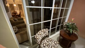 Ein Wohnzimmer mit einer Schiebetür zu einem Balkon. Es gibt eine Sofagarnitur, einen gemusterten Hocker und einen kleinen Tisch mit einer Pflanze.