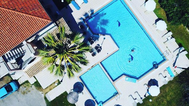 An aerial view of a hotel with a large rectangular swimming pool, palm trees, and lounge chairs.