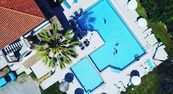 Vista aérea de un hotel con una amplia piscina rectangular, palmeras y sillas de descanso.