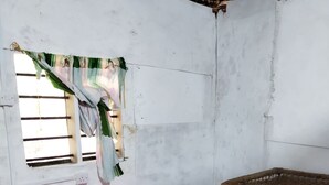 Una stanza con un tavolo coperto da una tovaglia rossa, una sedia in vimini, una finestra con grate e una tenda, e un muro bianco segnato dal tempo.
