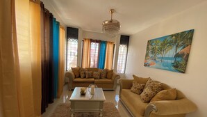 Uma sala de estar com dois sofás, uma mesa de centro, um candelabro e um quadro de uma paisagem tropical.