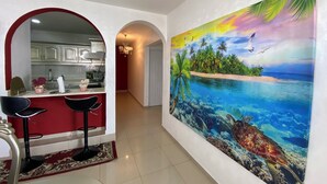 Um corredor com cozinha, área de refeições com duas cadeiras e um grande mural tropical na parede.