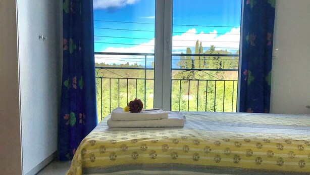 A bedroom with a bed, a window with a view of the sea, and a balcony with a railing. - Room-banos Apartments And Swimming Pool (Corfu)
