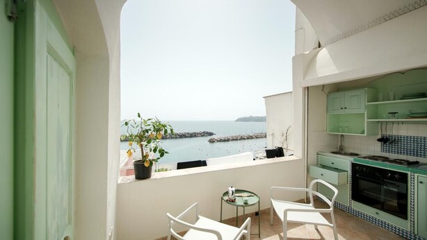 Un balcón con vistas al mar, una pequeña mesa, dos sillas y una cocina con armarios verdes.
