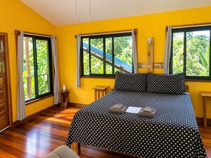 Une chambre avec un lit, un plancher en bois, deux fenêtres avec des rideaux et une vue sur la verdure à l’extérieur.