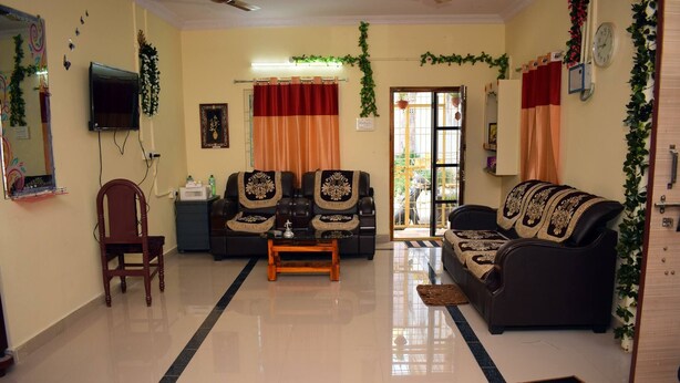 A well-lit living room with a sofa set, a wooden chair, a coffee table, and a television mounted on the wall.