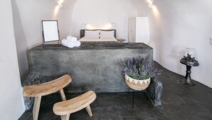 A bedroom with a bed, a bedside table, a chair, a potted plant, and a wall-mounted light fixture. - Luxury Cave Villa With Outdoor Hot Tub With Sea Sunset View (Santorini)