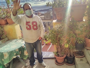 Una persona parada en un balcón con macetas, una mesa con mantel floral y vistas a edificios y agua al fondo.
