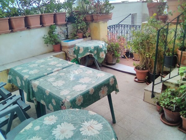 Uma área de pátio com duas mesas cobertas por toalhas florais, rodeada por vasos de plantas e um guarda-corpo de metal.