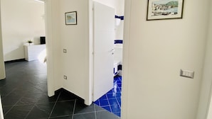 Ein Flur mit einer weißen Tür, die zu einem Badezimmer mit blauen Fliesen führt. An den Wänden befinden sich Bilder in Rahmen und auf einem Regal steht eine kleine Pflanze.