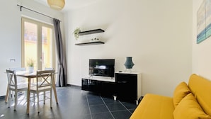 Ein modernes Wohnzimmer mit einem schwarzen Sofa, einem Esstisch mit Stühlen, einem Fernseher und einer Pendelleuchte.