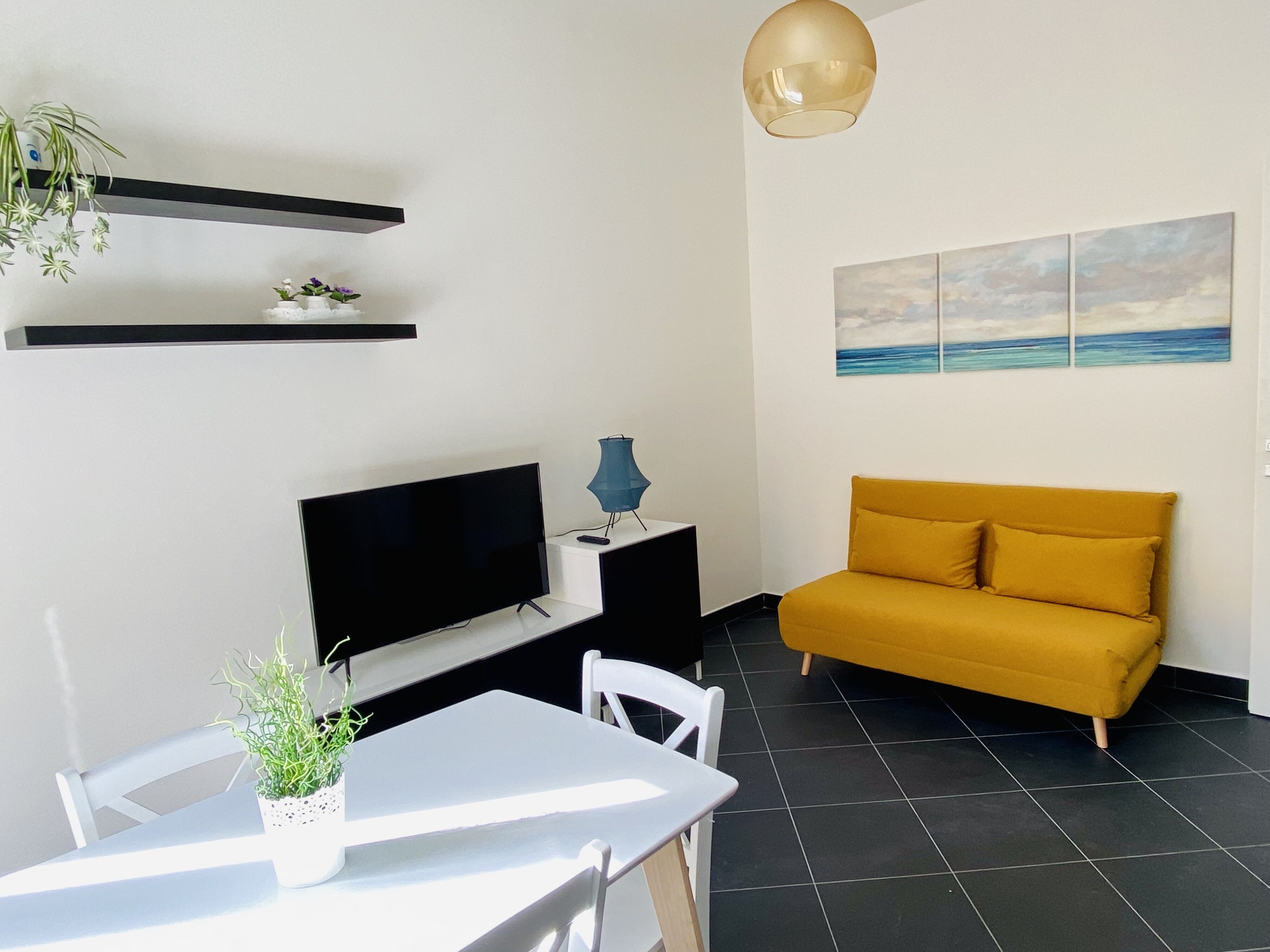 A modern living room with a flat-screen TV, a yellow sofa, a dining table with chairs, a potted plant, and a wall-mounted shelf with decorative items.