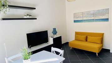 A modern living room with a flat-screen TV, a yellow sofa, a dining table with chairs, a potted plant, and a wall-mounted shelf with decorative items.