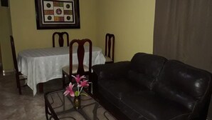A living room with a black leather sofa, a glass coffee table with a vase of flowers, a dining table with a white tablecloth, and a framed picture on the wall. - Apartahotel Next Nivel - Two Bedroom Apartment (Punta Cana)